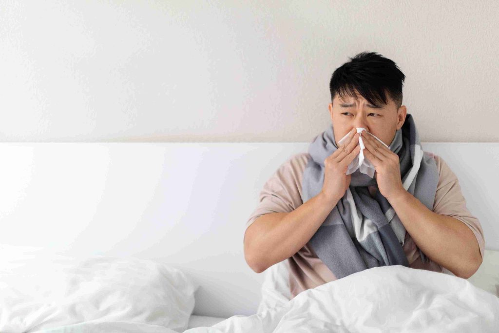 Man with flu, sitting in bed and covering his nose with a tissue.