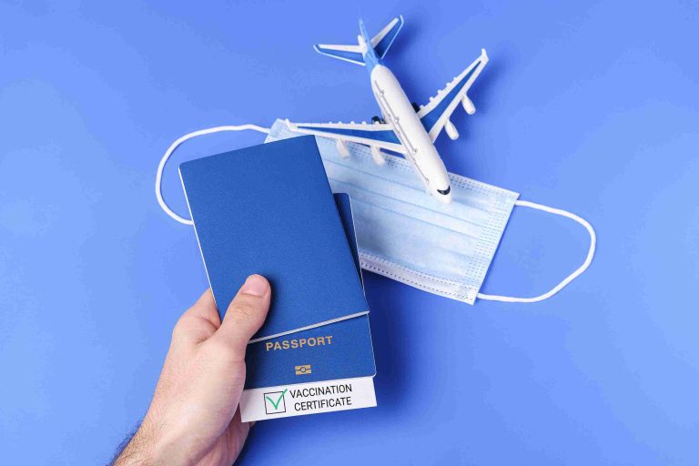 Image of a hand holding passports with vaccination pass next to a face mask and miniature airplane.